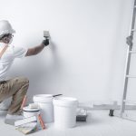 a man paints a white wall with a roller. Repair of the interior. Young male decorator painting a wall in the empty room, concept builder or painter in helmet with paint roller over the empty room.